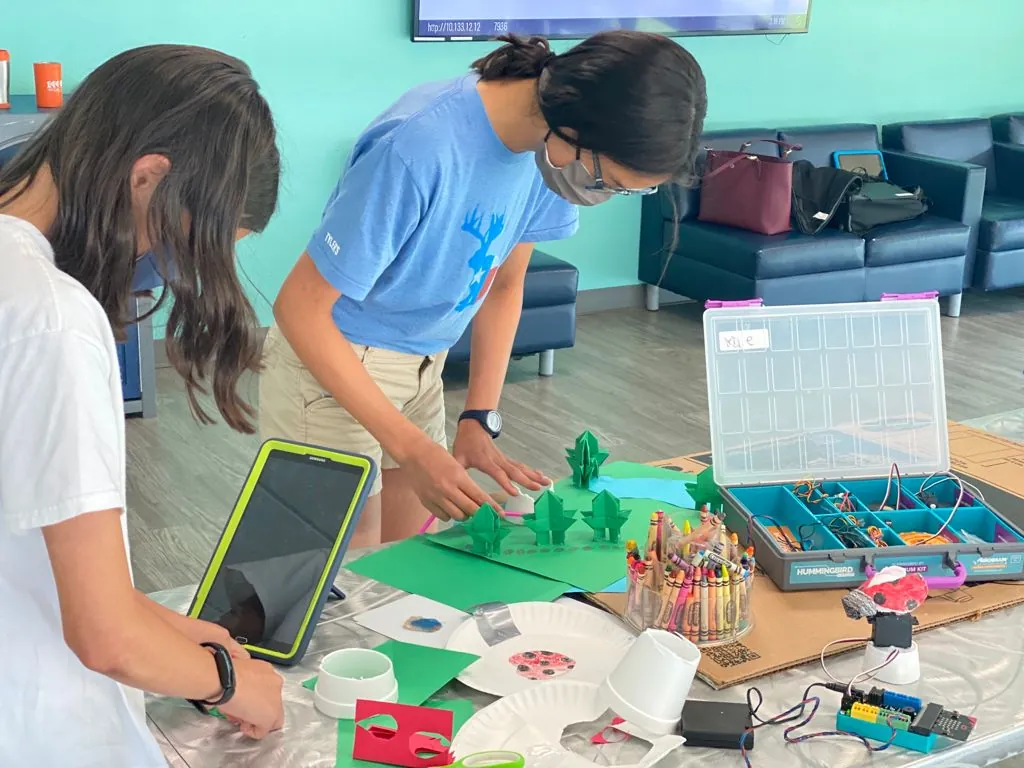 Two students building a robotics and design project with a tablet, craft materials and a robotics kit